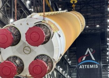 The four engine nozzles of NASA's orange Artemis III SLS core stage, covered in red caps, as cranes lift the stage inside the Vehicle Assembly Building under an Artemis banner