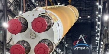 The four engine nozzles of NASA's orange Artemis III SLS core stage, covered in red caps, as cranes lift the stage inside the Vehicle Assembly Building under an Artemis banner