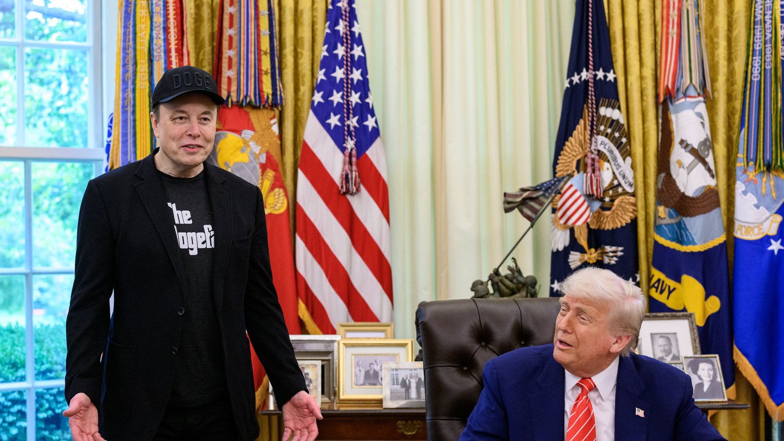Elon Musk stands and speaks beside President Donald Trump, seated at the Resolute Desk in the Oval Office