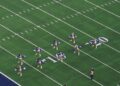 Dallas Cowboys Cheerleaders in blue and white uniforms with pom-poms perform in formation on the field at AT&T Stadium, seen from the upper deck
