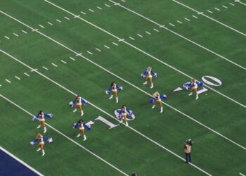 Dallas Cowboys Cheerleaders in blue and white uniforms with pom-poms perform in formation on the field at AT&T Stadium, seen from the upper deck