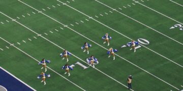 Dallas Cowboys Cheerleaders in blue and white uniforms with pom-poms perform in formation on the field at AT&T Stadium, seen from the upper deck