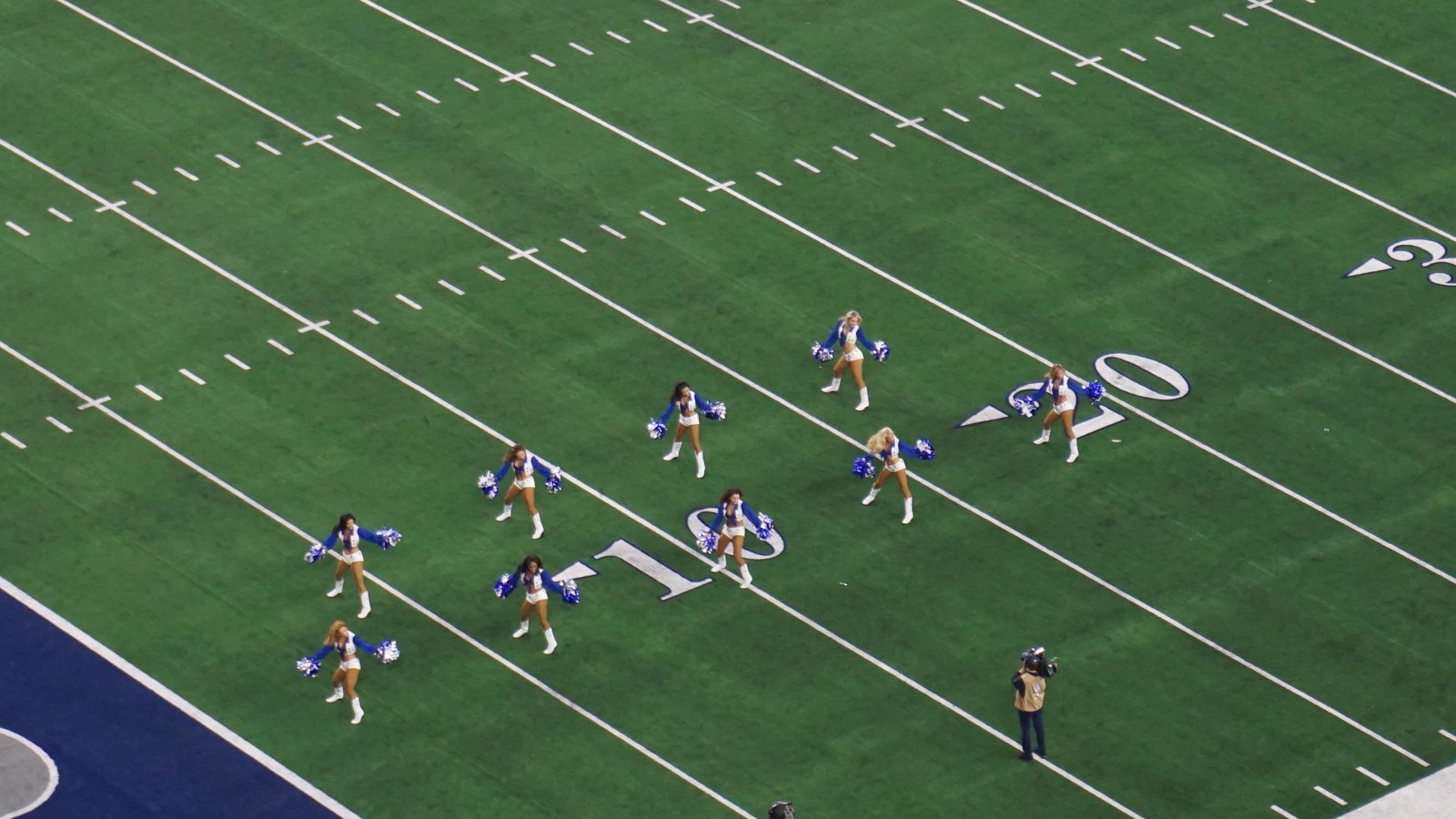 Dallas Cowboys Cheerleaders in blue and white uniforms with pom-poms perform in formation on the field at AT&T Stadium, seen from the upper deck