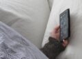 A person napping in bed under a checked blanket, one hand holding an e-reader against a white pillow