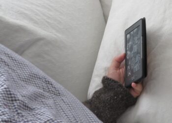 A person napping in bed under a checked blanket, one hand holding an e-reader against a white pillow