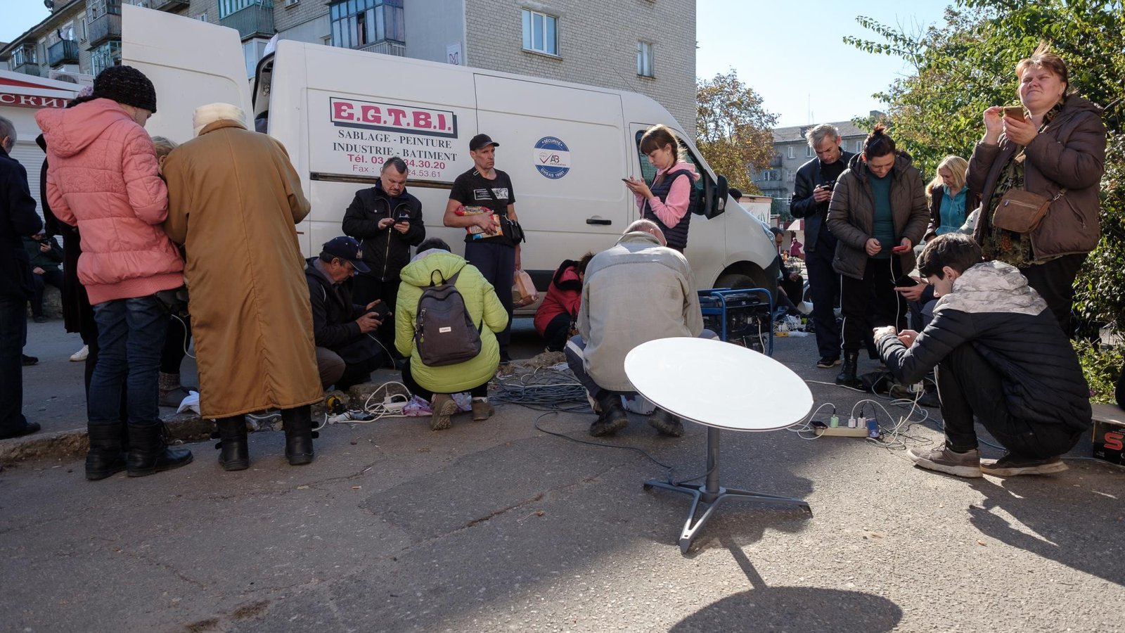 Residents of Kupiansk, Ukraine, charge and use their phones around a Starlink satellite dish set up on a street beside a white van