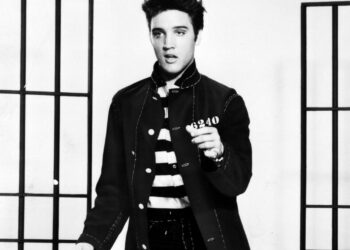 Elvis Presley in a dark jacket over a striped shirt, standing between prop jail bars in a black-and-white 1957 publicity photo for Jailhouse Rock