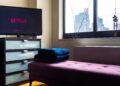 A TV showing the Netflix logo on a dresser in an empty living room beside a purple bench and a window overlooking city buildings