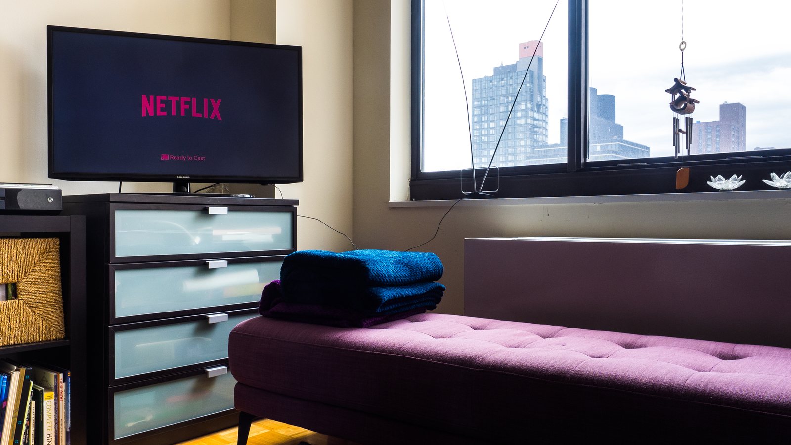 A TV showing the Netflix logo on a dresser in an empty living room beside a purple bench and a window overlooking city buildings