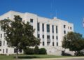The white stone Brazoria County Courthouse in Angleton, Texas, behind trees and a lawn under a blue sky