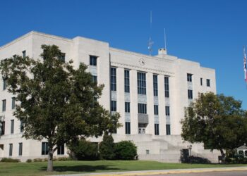 The white stone Brazoria County Courthouse in Angleton, Texas, behind trees and a lawn under a blue sky