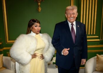 Nicki Minaj, in a white fur coat and yellow dress, stands beside President Donald Trump, who gives a thumbs-up, in a green-panelled room