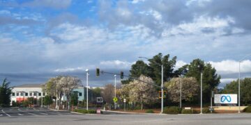 Meta's 1 Hacker Way headquarters sign beside a road intersection in Menlo Park, California, under a cloudy sky