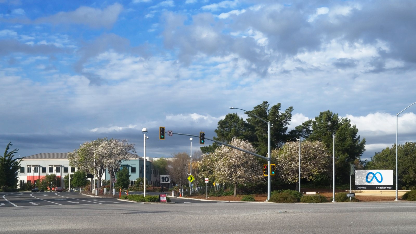 Meta's 1 Hacker Way headquarters sign beside a road intersection in Menlo Park, California, under a cloudy sky