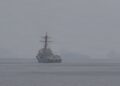 A US Navy guided-missile destroyer on hazy water in the Strait of Hormuz, with a helicopter overhead and rocky islands behind
