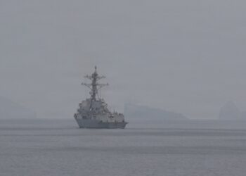 A US Navy guided-missile destroyer on hazy water in the Strait of Hormuz, with a helicopter overhead and rocky islands behind