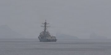 A US Navy guided-missile destroyer on hazy water in the Strait of Hormuz, with a helicopter overhead and rocky islands behind
